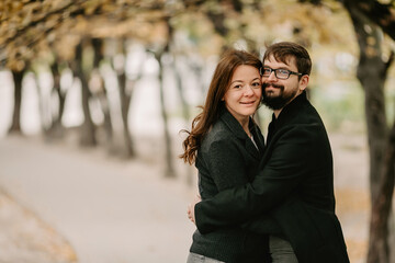 Fototapeta premium Portrait of loving couple cuddling on alley during autumn weather. Happy man with beard and eyeglasses, hugging with his girlfriend, while standing in park with yellowed leaves and looking at camera