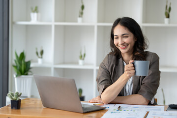 Asian businesswoman holding a coffee mug with a smile and working on a laptop computer with chart data The graph shows the results of the real estate business finance.