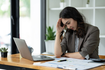 Tired stressed Asian businesswoman working on a laptop while sitting at a desk at the office and holding hands on temples. Migraine symptoms while calculating business income profit margin.