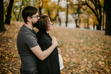 Side view of young family cuddling on alley. Loving husband, embracing his wife pregnant wife and warming her with jacket, while enjoying nature in park with fallen leaves