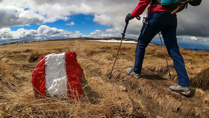 Woman walking next to path mark with Austrian flag painted on rock on remote alpine meadow near...