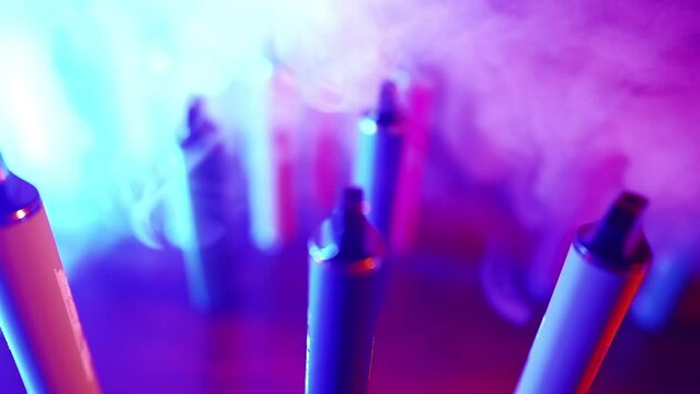 Lots of E-cigarettes and Vapes in Neon Lighting and Smoke. Concept of Bad Habits. Modern Alternative to Tobacco Products. Nicotine Addiction. Slow Motion.