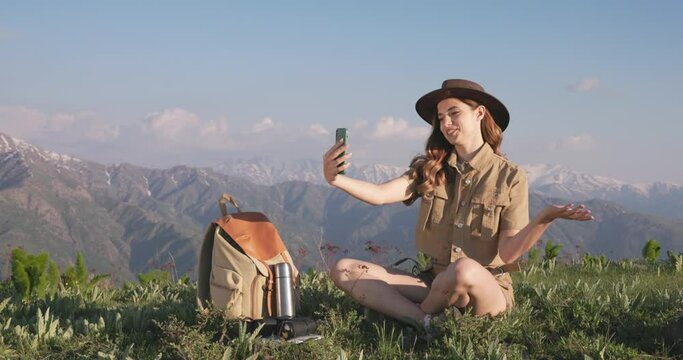 A woman hiker records video by smartphone for family and friends from her trip to mountains. Against background of beautiful mountains, millennial woman hiker smiles, shoots video and takes selfie