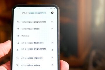 Close up of mobile phone screen with search "Will AI replace programmers" and list of suggested searches below. People searches on AI, concept of fear about AI replacing jobs. Social issue.