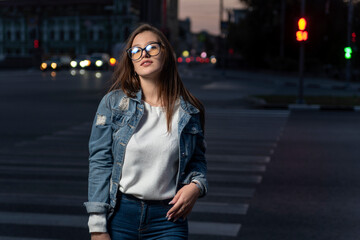 Portrait of brunette girl in youth clothes on evening city background. Stylish young woman in glasses on city street