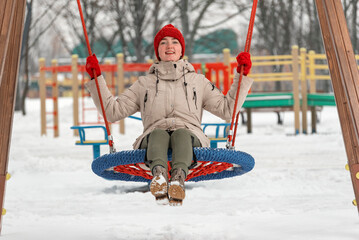 Laughing girl in red knitted hat cheerfully rides on swing on playground. Young woman has fun in winter in outdoors
