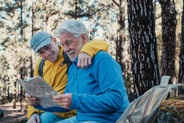 Fototapeta na wymiar Smiling caucasian couple of senior grandfather and young grandson hiking together in the woods resting looking at the map. Old and new generation sharing the passion for nature and healthy lifestyle.