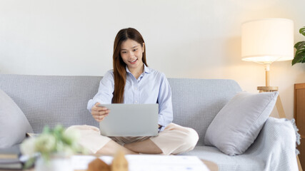 Obraz na płótnie Canvas Woman using laptop to work or do homework at home with smiling face in her living room, Creating happiness at work with a smile, Live performance or video call with laptop, Work from home.