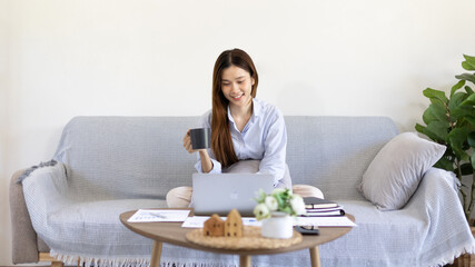 Obraz na płótnie Canvas Beautiful Asian woman working on laptop and sipping coffee with smiling face in her home, Creating happiness at work with a smile, Freelancer working at home happily, Work from home.