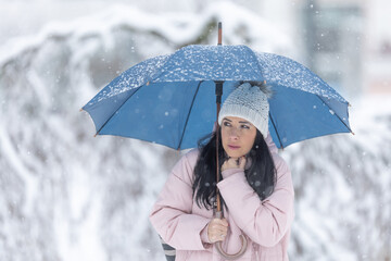 A frozen young woman shakes from winter under an umbrella during strong winds and snowfall