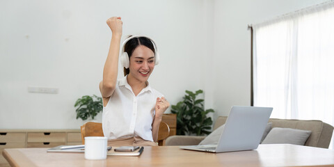 Asian teenage girl wearing headphones listening music while studying online on laptop, happy female student celebrating success sitting on floor in living room at home