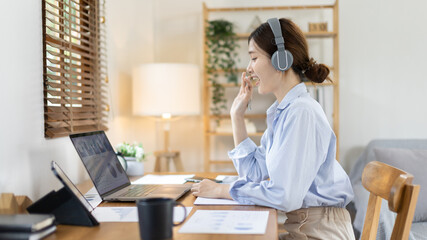Asian female college student wearing headphones watching live performance or video call teacher teaching on laptop, Conversations with teachers and classmates, Online learning, Study at your own home.