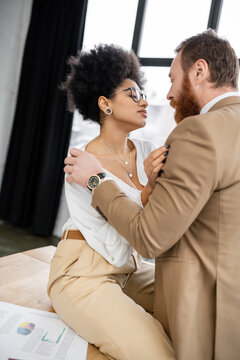 bearded man touching shirt while undressing seductive african american woman in office.