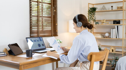 Asian female college student wearing headphones watching live performance or video call teacher teaching on laptop, Conversations with teachers and classmates, Online learning, Study at your own home.
