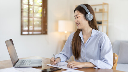 Asian female college student wearing headphones watching live performance or video call teacher teaching on laptop, Conversations with teachers and classmates, Online learning, Study at your own home.
