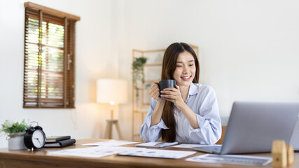 Beautiful Asian woman working on laptop and sipping coffee with smiling face in her home, Creating happiness at work with a smile, Freelancer working at home happily, Work from home.