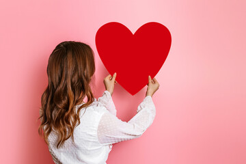 Girl hangs a big paper heart on a pink wall back view. Valentine's Day concept