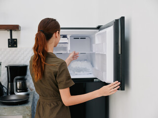 A woman in the kitchen of her home opened an empty freezer with ice inside, home refrigerator, defrosted, view from the back.