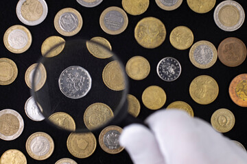 Collector or expert looking at coins in magnifying glass, top view of coin collection on black