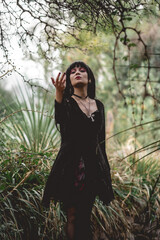 Young and skinny white goth girl with long black hair and black dress and pantyhose between plants in the forest