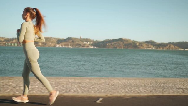 Confident fit female jogging by embankment area coastal city in beautiful sunny day do fitness training for cardio health. Tracking shot. Slow motion. Handheld. Athletic woman running, morning run