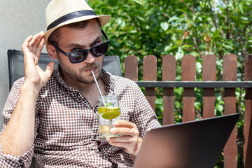 Handsome man with sunglasses and straw hat drinking fresh water with lime while sitting on terrace in summer using his laptop.