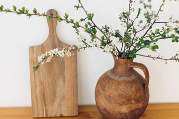 Blooming cherry branches in old vase and wooden board on table against white wall. Spring flowers...