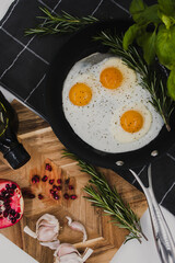 black frying pan on a black towel with three fried egg yolks in it next to a wooden cutting board with a red pomegranate on it.