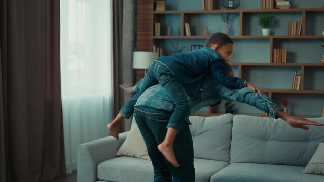 Happy African American father dad man piggyback riding on back little 5s preschool son boy child kid play airplane. Cheerful family playing together in cozy living room at home flying game playtime