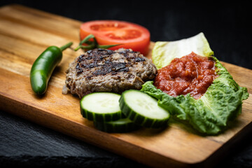 Homemade traditional Bulgarian grilled ground meat patty called kufte on wood surface decorated with dipping spread lutenica placed on lettuce lear next to tomato, cucumber and chili pepper