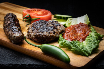 Bulgarian grilled meat patties on wooden board next to tomato and cucumber slices, jalapeño pepper and dipping sauce called lyutenica