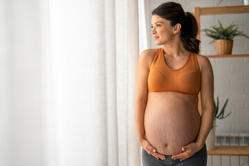 Beautiful pregnant woman touching her belly, standing by the window with the white curtains at home during the day. - obrazy, fototapety, plakaty