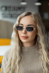 Naklejka premium Fashion female portrait of a beautiful young woman with vintage sunglasses with a curly blond hairstyle in a stylish beige sweater sits in a cafe