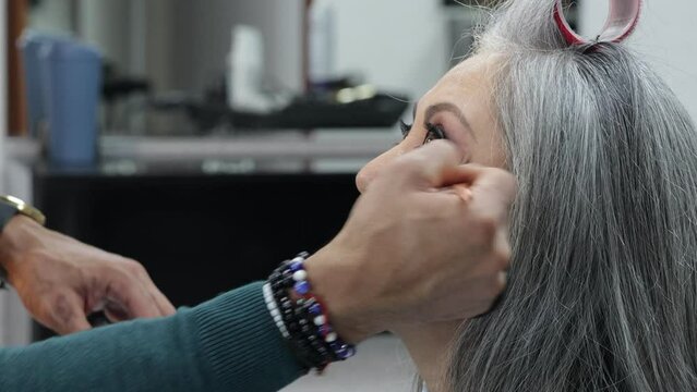 Professional makeup session on a senior model, applying makeup to her face, long gray hair with a hair curlers, tanned skin, serious expression. Fashion and beauty concept