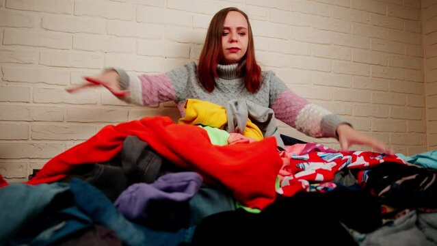Young unhappy woman sitting in pile of things on floor in room. Anxious female throwing scattered belongings aside.