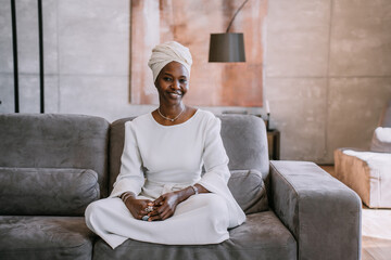 Satisfied African girl in white turban and traditional white dress sitting on couch looks at camera...