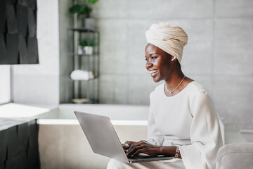 Laughing African young woman in white turban sitting at home with laptop using earphones, typing,...