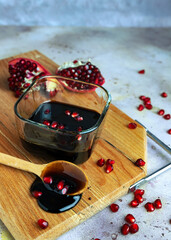 Natural organic pomegranate sause in glass cup, cut pomegranate fruit and grains on wooden cutting board on gray background. Selective focus. Blurred background.