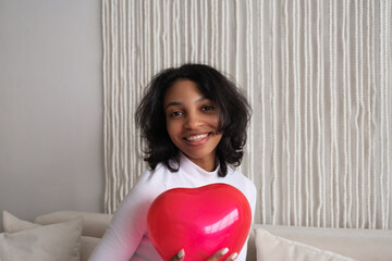 Closeup portrait of Young african american woman holding red heart shaped balloon sitting on sofa at home. Celebrating valentines,love day, women's day, birthday.