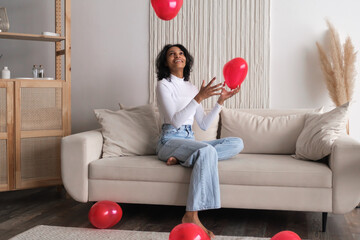 happy african american woman plays with red heart shaped balloons sitting on sofa at home. Celebrating valentines day concept