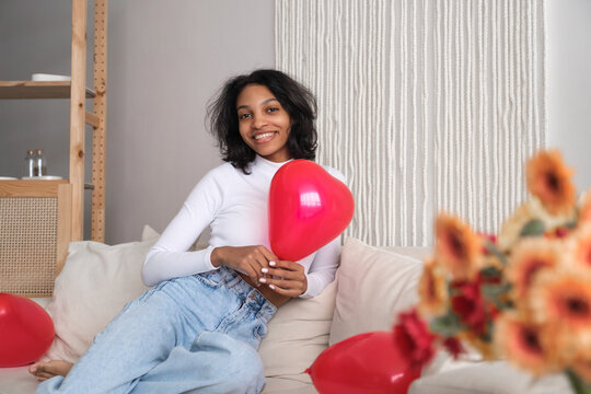 Young african american woman holding red heart shaped balloon sitting on sofa at home. Celebrating valentines,love day, women's day, birthday. Copy space banner - Powered by Adobe