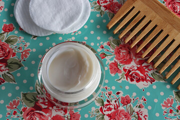 close up view of face cream, wooden hair comb and makeup remover pads on green floral background