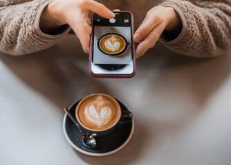 Young woman in a fluffy sweater keeps a smartphone in hands and makes a photo of a cup of coffee with heart shaped latte art foam. Close up cup of coffee with cream in coffee shop.