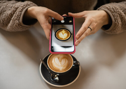 Young woman in a fluffy sweater keeps a smartphone in hands and makes a photo of a cup of coffee with heart shaped latte art foam. Close up cup of coffee with cream in coffee shop.