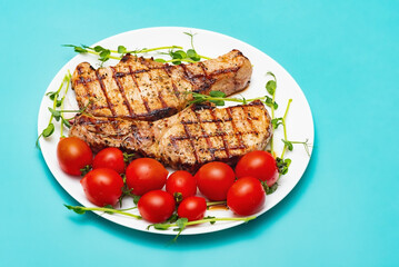 Two steaks on a white plate with tomatoes and greens on a turquoise background. Fried meat.