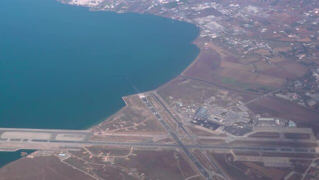 Flying above runway extended to sea, looking to the right of the frame. Passenger window view aboard airplane cruising at low altitude above SKG airport Thessaloniki, Greece.