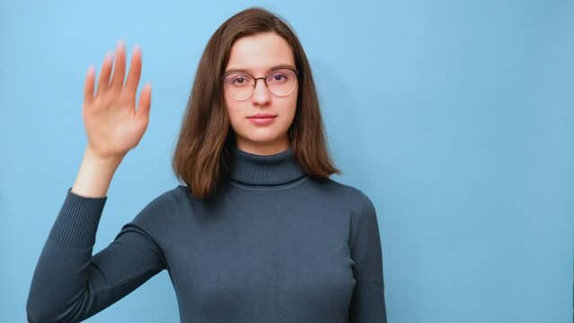 Portrait of a female student in glasses and a sweater waving goodbye sign on a blue background, 4K. Bye-bye. See you soon. Nonverbal communication