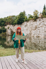 Fototapeta na wymiar Stylish hipster woman holding paper map, wearing backpack and red hat looking at mountain view while relaxing in nature. Travel and wanderlust concept. Amazing chill moment. Full body photo