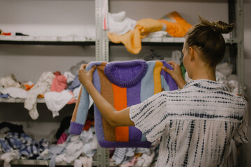 A young woman holding a multicolored purple striped wool knit children's sweater. Person picks out...