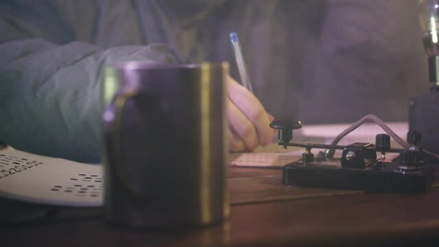 Man taps with finger on telegraph and writes notes in diary. Person communicates in Morse code sitting at table near mug with steam closeup